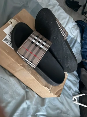Svarta sandaler med rutigt band - Säljer ett par svarta sandaler med breda band i klassiskt beige, svart, vitt och rött rutmönster. Sandalerna har en enkel slip-in design och är tillverkade i syntetmaterial med räfflad sula för bra grepp. Perfekta för en avslappnad och trendig stil. KAN RENA DOM ÅT DIG!! BARA SÄG TILL