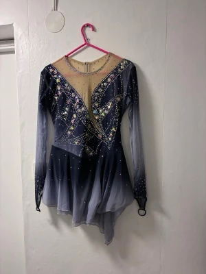Blå konståkningsklänning med strass - Säljer en snygg konståkningsklänning i blått med transparent mesh upptill och långa ärmar. Klänningen har massor av glittrande strass och pärlor i olika färger, samt en asymmetrisk kjol. Materialet är stretchigt och passar perfekt för isen. Den sitter bra på mig som är 162 cm lång och har ett stort höftmått. Använd 2 gånger, inga skador på.