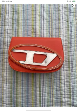 Orange handväska med stor metallogo - Cool orange handväska i slätt material med en stor, silverfärgad metallogo på framsidan. Väskan har en rundad form och lockstängning. Perfekt statement piece för dig som vill sticka ut.