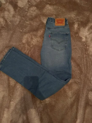 Blå Levi's raka jeans - Säljer ett par klassiska blå Levi's jeans med raka ben och fem fickor. Jeansen har en snygg tvättad look, Levi's ikoniska läderpatch bak och röda etiketten på bakfickan. Tillverkade i slitstark denim, perfekta för en avslappnad stil.