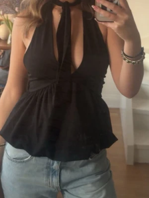 Svart halterneck topp med knyt (Bershka)  - Säljer en svart halterneck topp med djup v-ringning och knyt i nacken. Toppen har en figurnära passform upptill och faller ut nedtill för en snygg siluett. Perfekt till jeans eller kjol för en trendig look. 
