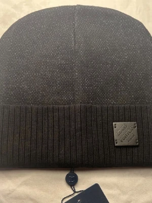 Svart mössa  - Snygg svart mössa  med diskret glittrande material och ribbad uppvikt kant. Märkets logga sitter på en svart patch framtill. Perfekt accessoar för dig som vill ha en exklusiv och stilren look.