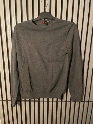 Mörkgrå basic tröja - En enkel och stilren mörkgrå tröja med rund halsringning och långa ärmar. Tröjan har ribbade muddar vid ärmslut och nederkant, perfekt för en clean och avslappnad look. Materialet känns mjukt och bekvämt mot huden.