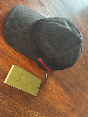 Svart Gucci keps med monogram - Snygg svart keps från Gucci med diskret GG-monogram över hela kepsen och klassiska gröna och röda ränder på sidan. Justerbar rem bak och material i canvas ger en exklusiv känsla. Perfekt accessoar för dig som vill sticka ut med stil.