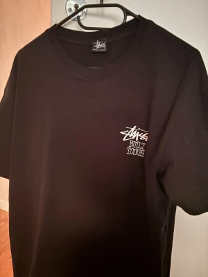 Svart Stüssy t-shirt med björntryck - Svart t-shirt från Stüssy med stort tryck av en nallebjörn och texten 'Built Tough' på ryggen. Framtill finns en mindre Stüssy-logga med samma text. Klassisk passform och rund hals. Perfekt för dig som gillar streetwear och unika prints.