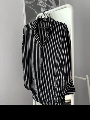Svartvitrandig skjorta med knappar - Snygg skjorta med svarta och vita vertikala ränder och klassisk krage. Skjortan har långa ärmar som kan rullas upp och fästas med knapp. Perfekt till jeans eller kjol för en trendig look.