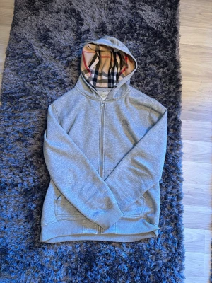 Burberry zip hoodie - Snygg grå zip-hoodie med huva och ikoniskt beige, svart och rött rutigt foder på insidan av huvan och hela insidan. Hoodien har fickor framtill och ribbade muddar vid ärmar och nederkant. Perfekt för dig som gillar stilrena detaljer och klassisk design.