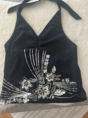 Svart halterneck topp med paljetter - Snygg svart halterneck topp med silverfärgade paljetter och blommigt mönster framtill. Toppen har knyt i nacken och är ärmlös med en lätt v-ringad urringning. Perfekt för dig som vill sticka ut lite extra.