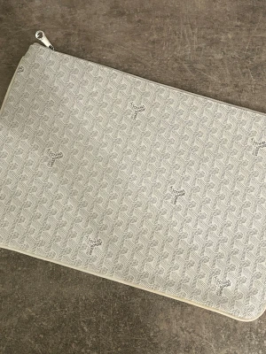 Beige kuvertväska från Goyard - Stilren beige kuvertväska från Goyard med ikoniskt mönster och diskret logga. Väskan har dragkedja upptill och är tillverkad i canvas med klassiskt Goyard-tryck. Perfekt för att bära det viktigaste med stil.