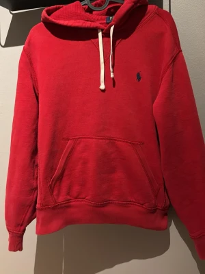 Röd hoodie från Polo Ralph Lauren - Röd hoodie med huva och vita dragsnören från Polo Ralph Lauren. Klassisk känguruficka framtill och broderad logga i mörkblått på bröstet. Tillverkad i mjukt bomullsmaterial och har ribbade muddar vid ärmar och nederkant.