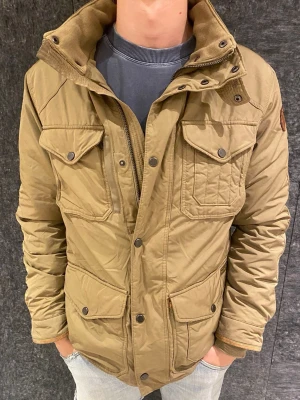 Polo Ralph lauren jacka  - Snygg beige parkajacka med flera stora fickor och tryckknappar. Jackan har hög krage, quiltade detaljer och patch på ärmen. Tillverkad i slitstarkt material, perfekt för kyligare dagar. Stängs med både dragkedja och knappar.