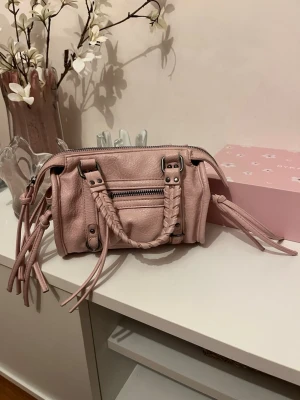 Ljusrosa handväska med flätade handtag - Söt ljusrosa handväska i mjukt skinnliknande material med flätade handtag och dekorativa dragkedjor. Väskan har silverfärgade detaljer och långa tofsar på sidorna för en trendig look. Perfekt storlek för det viktigaste och ger en gullig touch till din outfit.