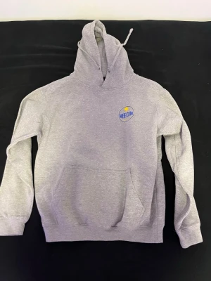 Grå hoodie med tryck från Wearclan - Snygg grå hoodie från Wearclan med liten logga i blått och gult på bröstet. Klassisk modell med huva, känguruficka och ribbade muddar. Perfekt för en chill och avslappnad stil. Materialet känns mjukt och bekvämt.