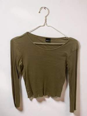 grön ribbad långärmad topp - Snygg olivgrön långärmad topp med ribbad struktur och croppad passform. Rund halsringning och vågig kant nedtill ger en trendig look. Perfekt att matcha med höga jeans eller kjol.