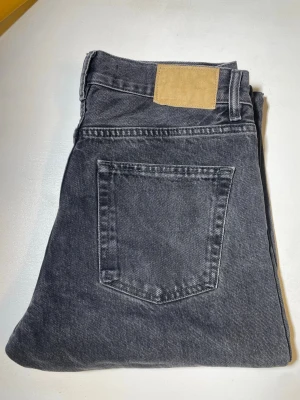 Svarta raka jeans med klassisk passform - Säljer ett par grå jeans med rak passform och klassisk femficksdesign. Jeansen har en beige patch bak i midjan och är tillverkade i mjukt denimtyg. Perfekta för en avslappnad och stilren look.
