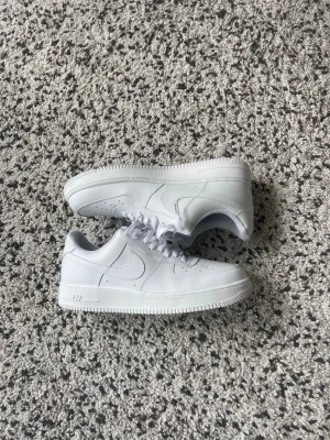 Nike Air Force 1 vita sneakers - Klassiska helvita Nike Air Force 1 sneakers i lågt utförande. Skorna har en stilren design med perforerade detaljer på tån, vit sula och snörning. Tillverkade i läder med diskret Nike-logga på sidan och tungan. Perfekta för en clean och tidlös look.