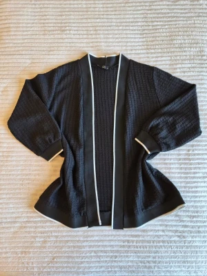 Svart kofta från ON Fashion stl M - Stilren svart kofta från ON Fashion med strukturstickat tyg och breda, kontrasterande vita kanter. Koftan har vida ärmar och öppen front, vilket ger en avslappnad men trendig look. Perfekt att slänga över en t-shirt eller topp för extra stil.