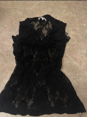 Svart spetslinne med volanger - Snyggt svart linne i genombruten spets med volangdetaljer vid bröstet. Figurnära modell med ärmlös design och rundad hals. Perfekt för dig som vill ha en trendig och feminin topp med extra detaljer.