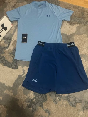 Blått träningsset från Under Armour - Snyggt träningsset från Under Armour med en ljusblå t-shirt och mörkblå shorts. T-shirten har kort ärm och rund hals, medan shortsen har elastisk midja och logga på benet. Perfekt för gym eller löpning. Tillverkad i funktionsmaterial för bästa komfort.