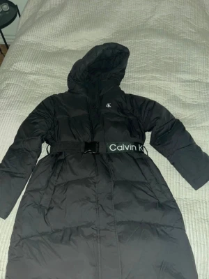 Svart lång dunjacka Calvin Klein - Snygg svart lång dunjacka från Calvin Klein med huva och bälte i midjan där det står Calvin Klein i vitt. Jackan har puffig design och diskret CK-logga på bröstet. Perfekt för kalla vinterdagar och riktigt trendig look. Jackan har ett knappt synligt hål vid fickan men annars i fint skick 