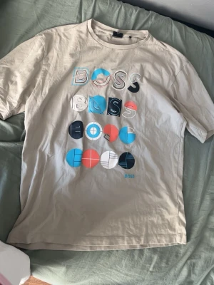 Beige BOSS t-shirt med grafiskt tryck - Säljer en beige t-shirt från BOSS i storlek M. T-shirten har ett färgglatt grafiskt tryck framtill med bokstäver och cirklar i blått, svart, vitt och rött. Klassisk rund halsringning och korta ärmar. Perfekt för en avslappnad och trendig stil.