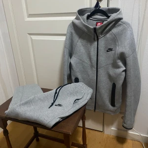 Nike Tech Tracksuit - Säljer en grå hoodie från Nike i storlek XS. Tröjan har huva, hel dragkedja framtill och svarta detaljer vid fickorna. Nike-loggan är broderad på bröstet. Materialet är mjukt och känns som bomull och polyester, perfekt för chill eller träning. Defekt på byxorna 