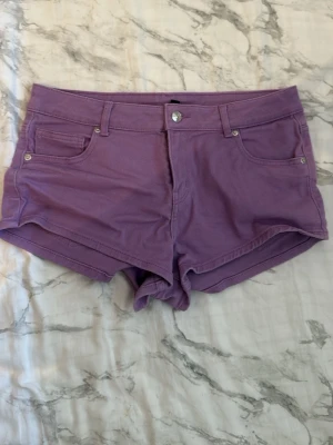 Lila shorts med fem fickor - Snygga lila shorts i klassisk femficksmodell med knapp och dragkedja framtill. Shortsen har bälteshällor och är tillverkade i ett mjukt bomullstyg. Perfekta för varma dagar och enkla att matcha med olika toppar.