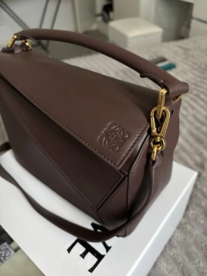 Brun geometrisk handväska i läder - Snygg brun handväska från Loewe i mjukt läder med geometrisk design och guldfärgade metalldetaljer. Väskan har både handtag och axelrem för olika stilar. Perfekt för dig som gillar stilrena och moderna accessoarer.