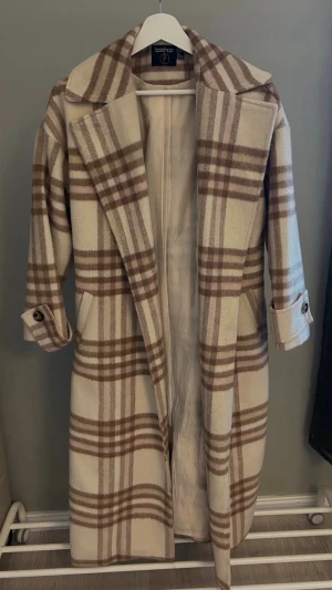 Beige-rutig kappa från Boohoo - Stilren lång kappa i beige med brunt rutmönster från Boohoo. Kappan har bred krage, knytskärp i midjan och dekorativa knappar vid ärmsluten. Perfekt för att lyfta din höst- eller vinterstil med en trendig touch.
