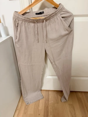 Beige linnebyxor med dragsko XS - Snygga beige linnebyxor från lager 157 i storlek XS. Byxorna har avslappnad passform, resårmidja med dragsko och praktiska sidofickor. Perfekta för en chill och stilren look. Tillverkade i ett lätt och luftigt linnematerial.
