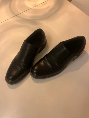 Svarta klassiska loafers i skinn - Säljer ett par svarta loafers i äkta skinn med slät ovandel och rundad tå. Skorna har diskreta sömmar och elastiska paneler på sidorna för enkel påtagning. Yttersulan är i gummi med cirkulära mönster för extra grepp.