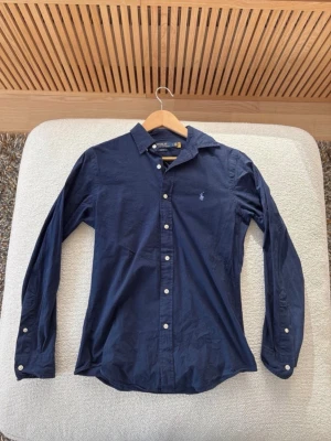 Mörkblå skjorta från Polo Ralph Lauren - Klassisk mörkblå skjorta från Polo Ralph Lauren med vit broderad logga på bröstet. Skjortan har långa ärmar, knappar framtill och vid manschetterna samt en tidlös, stilren design. Perfekt till jeans eller chinos för en clean look.