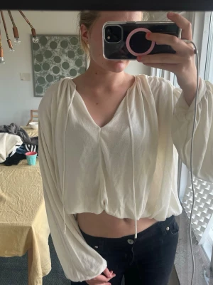 Vit croppad blus med v-ringning - Säljer en luftig vit blus med v-ringning och långa, vida ärmar. Blusen har dragsko i halsen och är croppad i modellen vilket ger en avslappnad och trendig look. Perfekt att matcha med höga jeans eller kjol. Helt oanvänd. 