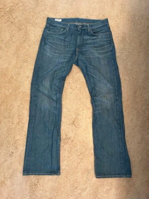 Levis 527 - Säljer ett par klassiska blå jeans med bootcut passform och fem fickor. Jeansen har normal midja, knappgylf och subtila slitningar på baksidan av knäna. Materialet är denim i bomull och färgen är mellanblå med lätt tvättad look.