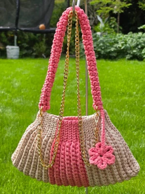 Beige och rosa virkad handväska - Unik handväska i beige och rosa, virkad i tjockt garn med guldfärgade kedjedetaljer. Väskan har en bred axelrem och en dekorativ virkad blomma i rosa. Perfekt för dig som gillar färg och handgjorda accessoarer.