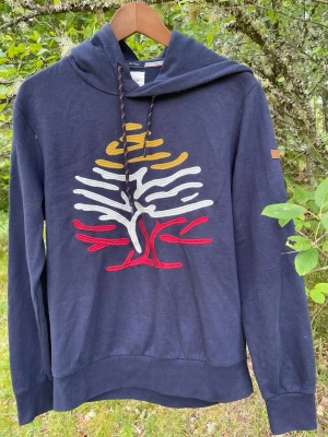 Mörkblå hoodie med broderat träd - Mörkblå hoodie från Springfield med ett stort broderat träd i vitt, gult och rött på bröstet. Hoodien har dragsko i huvan och ribbade muddar vid ärmslut och nederkant. Tillverkad i mjukt material för en skön känsla.