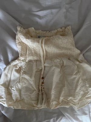 Krämvit kjol med snörning - Säljer ett par krämvita kjol  med smockdetaljer och volangkant. Kjolen  har snörning framtill med dekorativa träpärlor och är tillverkade i ett lätt, luftigt material som passar perfekt till varma dagar.