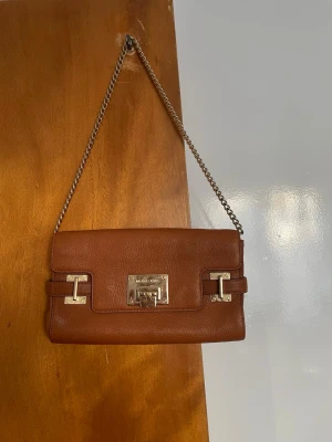 Brun axelremsväska Michael Kors - Snygg brun axelväska från Michael Kors i äkta läder med silverfärgade metalldetaljer och kedjerem. Väskan har ett stilrent rektangulärt lock med vridlås och diskret logga på framsidan. Perfekt för dig som gillar klassisk och trendig design. Axelrem: 67cm. 
