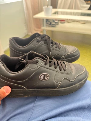  Champion - Säljer ett par svarta sneakers från Champion med lågt skaft och klassisk snörning. Skorna har en diskret logga på sidan och är tillverkade i syntetmaterial med gummisula. Perfekta för en clean och sportig look.