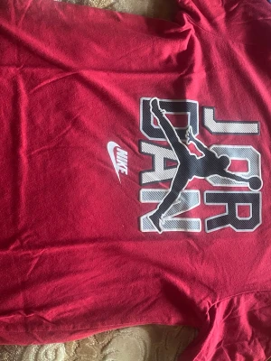 Röd Jordan t-shirt från Nike - Snygg röd t-shirt från Nike med stort Jordan-tryck och Jumpman-logga på bröstet. Klassisk passform och korta ärmar, perfekt för en sportig och avslappnad stil. Materialet känns mjukt och bekvämt mot huden.