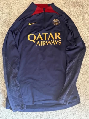 PSG träningströja i mörkblått - Mörkblå långärmad träningströja från Nike med PSG-logga och Qatar Airways-tryck i gult på bröstet. Tröjan har halv dragkedja, röda detaljer vid halsen och diskret mönster på ärmarna. Perfekt för fotbollsträning eller sportiga aktiviteter.