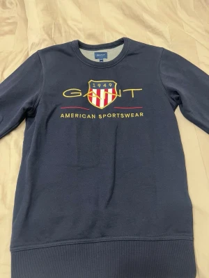Mörkblå sweatshirt från GANT XS - Mörkblå sweatshirt från GANT med broderad logga och sköld på bröstet. Texten 'American Sportswear' i gult och rött ger en sportig vibe. Rund halsringning och mjukt material, perfekt för en avslappnad stil.