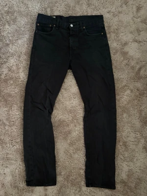 Svarta raka jeans - Säljer ett par svarta 501 jeans med rak passform och klassisk femficksdesign. Jeansen har knappgylf och bälteshällor. Materialet ser ut att vara bomullsdenim med en stilren look. Perfekta för dig som gillar en enkel och tidlös stil.