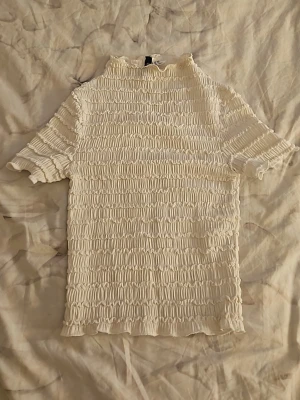 Vit ribbad topp från H&M Divided - Säljer en vit ribbad topp från H&M Divided i storlek XXS. Toppen har korta ärmar, hög hals och ett stretchigt, smockat tyg som ger en snygg och bekväm passform. Perfekt att styla med jeans eller kjol för en trendig look.