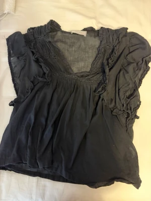 Svart neo Noir blus - Snygg svart blus med djup v-ringning och volangdetaljer längs ärmar och hals. Blusen har en lös passform och är tillverkad i ett tunt, mjukt tyg som ger ett luftigt intryck. Perfekt för dig som gillar en avslappnad men trendig stil.