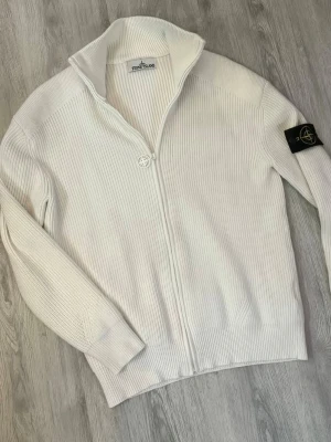 Vit stickad ziptröja från Stone Island - Vit ribbstickad ziptröja från Stone Island med hög krage och ikonisk logotyp-patch på vänster ärm. Tröjan har hel dragkedja framtill och långa ärmar med ribbade muddar. Perfekt för dig som gillar stilrena och klassiska plagg.