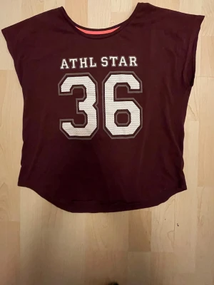Vinröd t-shirt med ATHL STAR-tryck - Snygg vinröd t-shirt med stort vitt ATHL STAR-tryck och siffran 36 framtill. T-shirten har en avslappnad passform och ärmlös design, perfekt för en sportig och trendig look. Tillverkad i mjuk bomull för skön känsla hela dagen.