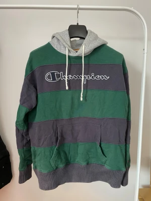 Randig Champion hoodie - Säljer en hoodie från Champion med breda gröna och blåa ränder. Stor broderad Champion-logga på bröstet och grå huva med vita snören. Känguruficka framtill och ribbade muddar. Perfekt för en avslappnad streetwear-look.
