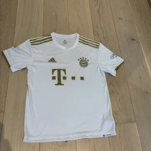 Vit Bayern München fotbollströja Adidas - Snygg vit Bayern München fotbollströja från Adidas med guldiga detaljer. Tröjan har korta ärmar, guldiga ränder på axlarna och klubbmärke samt sponsorlogga på bröstet. Tillverkad i lätt och ventilerande material, perfekt för match eller träning.