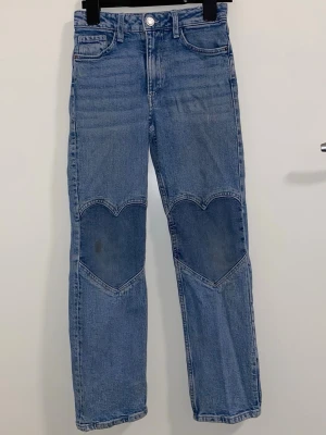 Blå jeans med hjärtlappar - Säljer ett par ljusblå raka jeans i denim med unika hjärtformade lappar på knäna. Klassisk femficksmodell och normal midja. Materialet är bomullsdenim och byxorna har en avslappnad passform som ger en trendig vibe.
